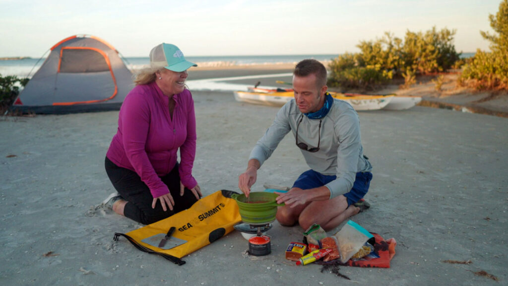 Chef Corso and Guide Misty Wells kneeling on the beach cooking a meal over a camp stove.